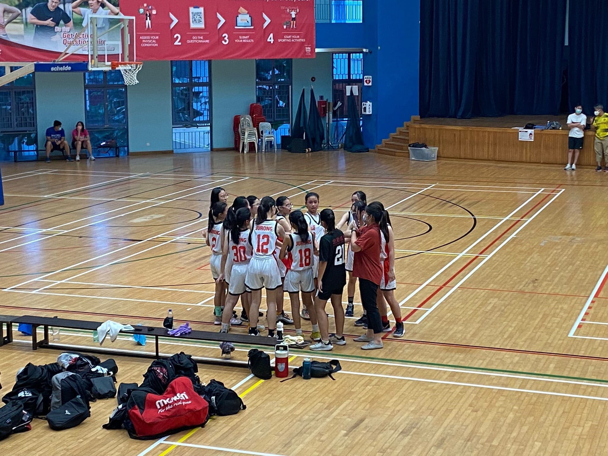 A girls basketball team in white Jurong uniforms huddles with a coach on an indoor wooden court.
